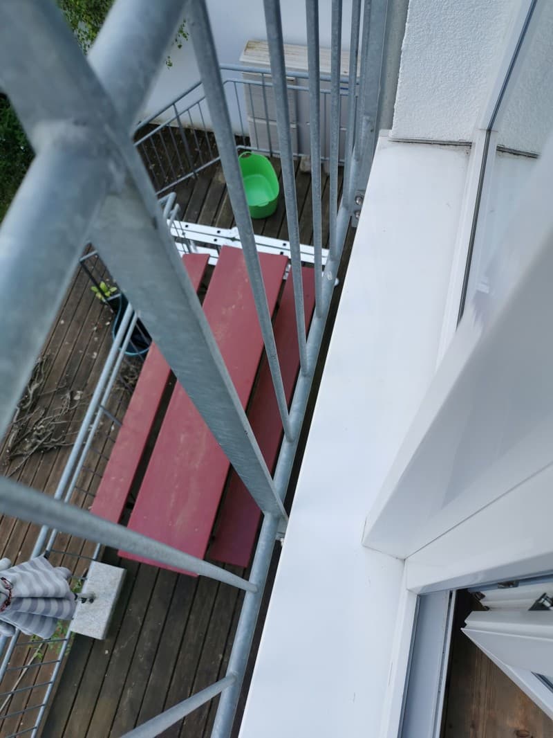 Ein Blick von oben auf einen Balkon mit einem grauen Metallgeländer, einem roten Tisch und einem grünen Eimer. Der Boden ist aus Holz.