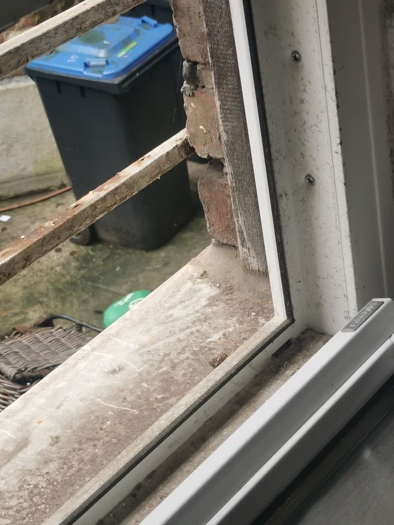 Nahaufnahme eines Fensterrahmens mit Blick auf Mauerwerk und eine blaue Mülltonne im Hintergrund. Der Fensterrahmen ist weiß und wirkt etwas schmutzig.