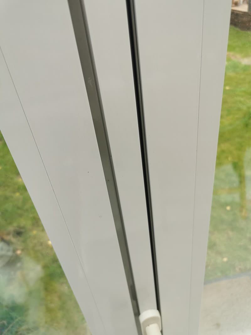 Eine Nahaufnahme eines weißen Fensterrahmens mit einem grauen Profil und einer schwarzen Dichtung. Der Hintergrund zeigt einen verschwommenen Blick auf einen Garten.