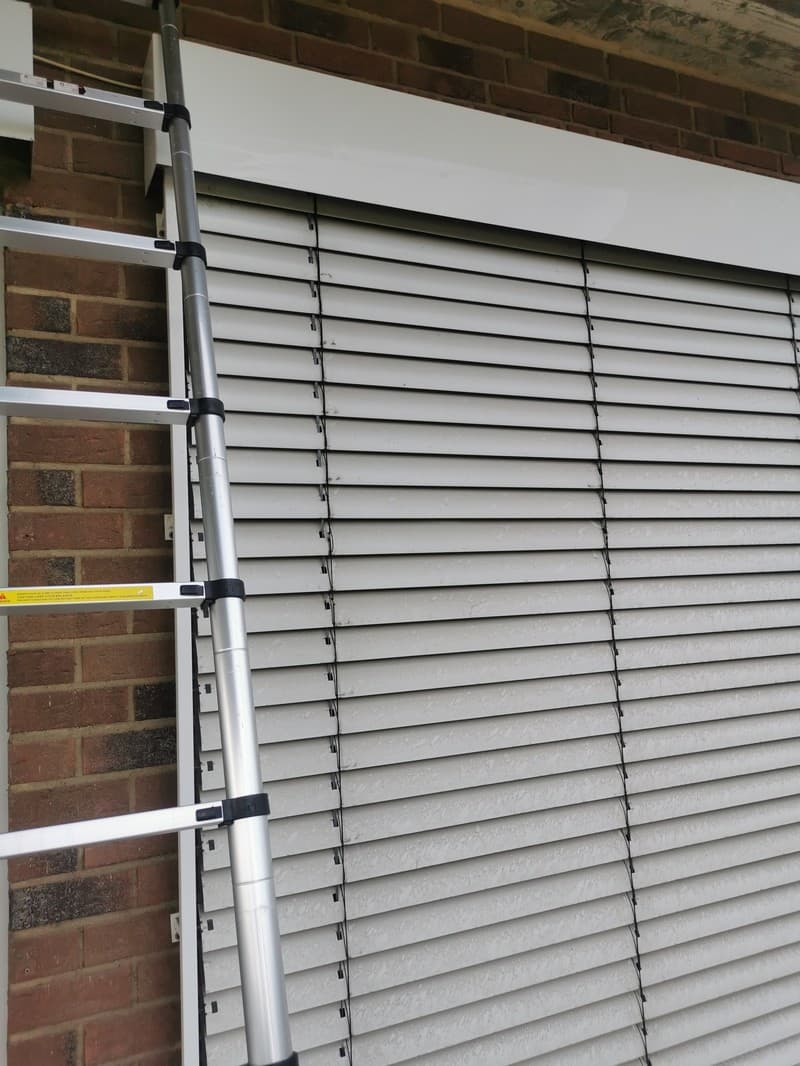 Eine silberne Leiter steht an einer Backsteinmauer neben einem Fenster mit geschlossenen Jalousien. Die Jalousien sind grau und horizontal ausgerichtet.
