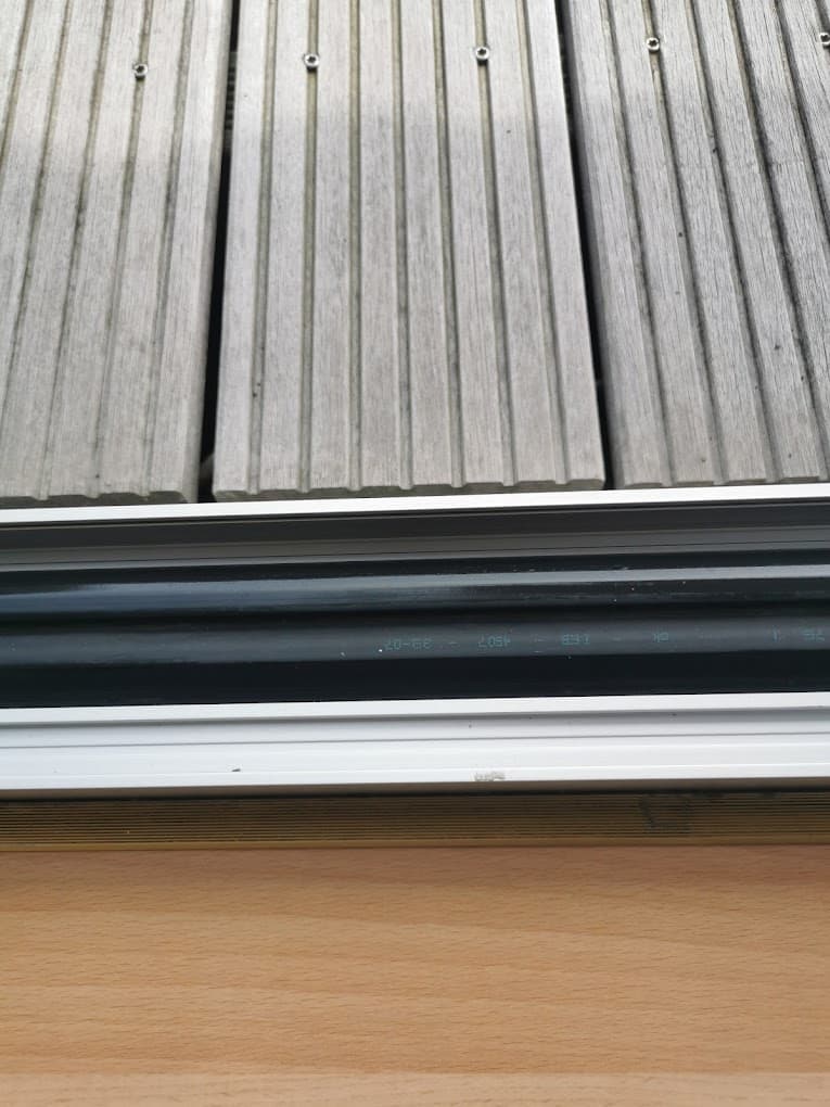 Nahaufnahme einer Terrasse mit Holzboden, einem weißen Fensterrahmen und einem schwarzen Kabel. Die Holzplanken der Terrasse sind grau und haben Rillen.